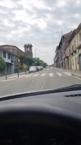 A dash cam view from inside a car shows a street in what appears to be a European town. The sky is overcast with clouds. On the left side of the street, there are buildings and parked cars. A distinctive building with a tower, possibly a church, is visible. On the right side of the street, there are more buildings and a sidewalk. The car is driving down the street, passing a crosswalk and several parked cars. A few cars are also driving on the road ahead. The road surface is asphalt, and there are white lines marking the lanes and the crosswalk. The overall scene suggests a typical day in a moderately populated urban area.