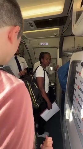 A medium shot captures the interior of an airplane aisle, focused on a person wearing a pink shirt viewed from behind. To the left, two individuals in white shirts are visible, one appearing to be a crew member with a dark tie. In the center of the aisle, a Black woman carrying a large black backpack with yellow accents and a clipboard stands facing right. Behind her, a man in a light-colored uniform, likely a pilot, can be seen looking down and gesturing with his hands. The overhead compartment and an "EXIT" sign are visible. The right side of the frame shows a portion of an airplane seat with blue fabric and part of a window with tape displaying "ISRAEL AIR" text. The lighting appears to be artificial, emanating from above.