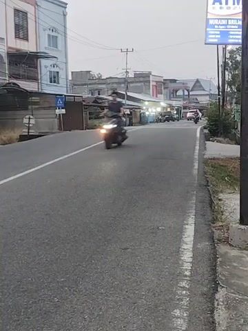 A person is riding a motorcycle down a paved road. The person is wearing a red shirt. The road is lined with buildings and shops. There is an ATM sign on the right side of the road. The sky is overcast. The motorcycle is moving from left to right in the frame.