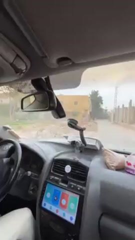 The video shows the view from inside a car as it drives down a narrow street. The interior of the car is visible, including the dashboard with a touchscreen displaying colorful icons, the steering wheel, and the rearview mirror. The driver's hand is briefly visible on the steering wheel. The street outside is lined with buildings and stone fences, with some vegetation visible. The sky is overcast. Someone in the car speaks in German. The car appears to be navigating a residential area.