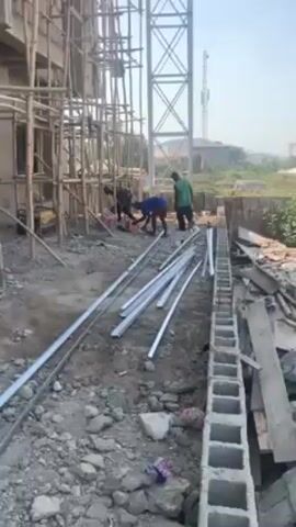 The video shows an outdoor construction site. A building is under construction, with bamboo scaffolding erected around its exterior.  A tall, white steel tower stands nearby. Several long, white metal pipes lie on the ground. Two men are working near the pipes and a cinder block wall.  One man, wearing a blue shirt and red shorts, appears to be operating a tool on the ground. The second man is wearing a green shirt and dark pants.  In the background, there are more buildings and greenery, under a clear, light blue sky. Construction debris is scattered around the work area.