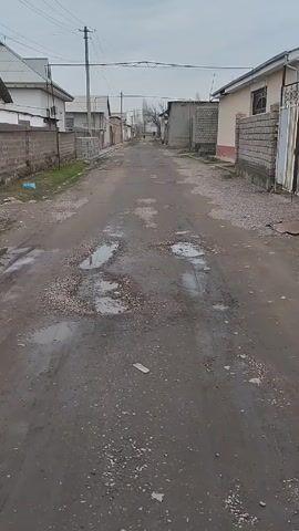 The video shows a deserted, unpaved road winding through a neighborhood. The road is heavily damaged with numerous potholes filled with water. The sky is overcast, creating a somber atmosphere. Houses and brick walls line both sides of the road. A utility pole stands near the beginning of the street, with wires stretching across the sky. In the distance, a figure can be seen walking away from the camera. The overall scene appears to be a quiet, impoverished area.