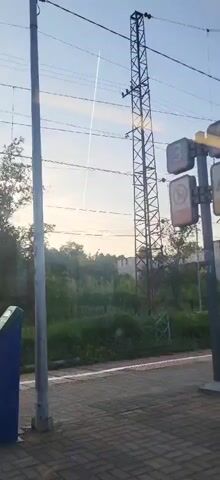 The video displays an outdoor scene, viewed from behind a window with visible reflections. A long, white contrail extends diagonally across a light blue sky from the upper left to the mid-right. In the midground, a tall, dark metal lattice pylon stands centrally, with several horizontal wires crossing the frame. To the right of the pylon, a signal post features at least three rectangular signs, one displaying the number "3" and another a circular prohibition symbol. A green, overgrown area with trees is visible behind these structures, and a light-colored building is faintly seen in the far background. In the foreground, a paved or brick platform or sidewalk is visible. A vertical gray pole stands on the left. No individuals are present in the frame.