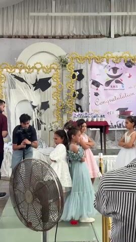 The video shows a graduation ceremony for young children. Several girls, dressed in formal dresses, stand in front of a decorated backdrop. Some are posing for photos, while others are looking at the camera. A man with a camera is filming the event. A large fan is visible in the foreground. The backdrop has graduation caps and a banner that says "Congratulations".