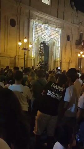 A nighttime shot shows a crowd of people gathered outside a building with ornate, illuminated arches over its entrance. The building appears to be a church or a public institution, with a light-colored facade and several windows visible. The arches are decorated with a pattern of many small lights. Streetlights also illuminate the scene. People in the crowd are mostly facing towards the entrance, with some individuals holding up phones, presumably to record the event. A person in the foreground is wearing a black t-shirt with the words "HAPPY TOGETHER" and smiley faces printed on it. The overall atmosphere appears festive, possibly indicating a religious procession, a festival, or a public celebration.