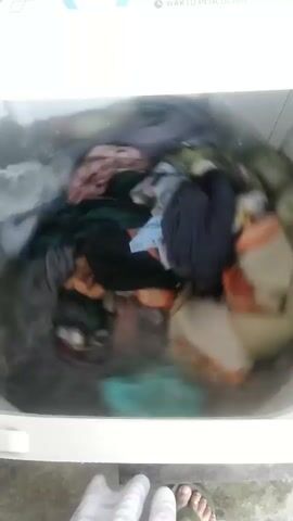 The video shows the inside of a washing machine filled with clothes and water. The clothes are a mix of colors and patterns, including dark blues, greens, oranges, and whites. The washing machine appears to be in operation, as the clothes are swirling around in the water. In the foreground, the feet of a person wearing flip-flops are visible, along with a portion of a light-colored towel or blanket. The floor beneath the washing machine is visible and appears to be concrete. The scene suggests a mundane domestic activity of laundry being done.