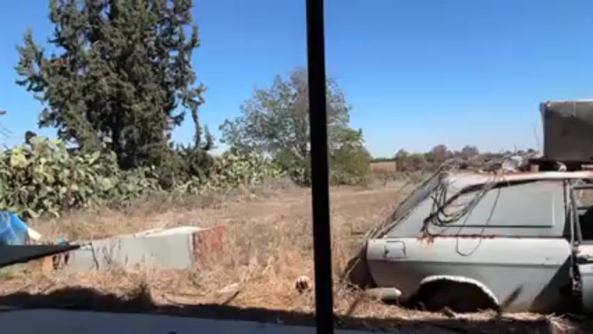 The video shows a dilapidated grey station wagon, likely abandoned in a field. The car has extensive rust and is missing windows. There's a wooden target mounted on the roof, surrounded by wires. The surrounding environment includes dry grass, trees, and cacti under a clear blue sky. The camera appears to be positioned behind a pole or structure, offering a partial view of the car.