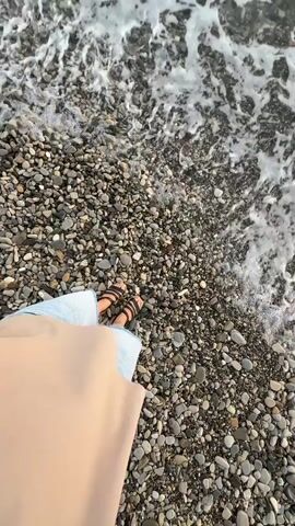 The video opens with a top-down view of a person's feet clad in sandals and light blue pants standing on a rocky shore. Gentle waves with white foam lap against the shore, momentarily covering the lower half of the person's feet and the surrounding pebbles. The camera slowly pans upwards, revealing the texture of the beach and the movement of the water. The person's hand, wearing a light-colored sleeve, is visible in the foreground, suggesting a first-person perspective. The environment is a beach with numerous small, grey, and brown pebbles. The lighting suggests it is daytime, possibly late afternoon or early morning due to the soft light.