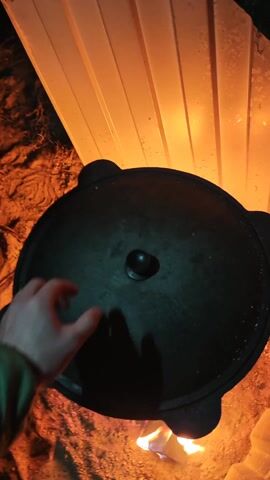 A high-angle shot depicts a hand removing the lid from a large, black cauldron situated over an open fire. Inside, a mixture of chicken and carrots simmers. The camera then shows a second hand using a fork to add chopped tomatoes from a white plate to the cauldron. The cauldron is placed on a three-legged stand directly over the flames, with the immediate environment consisting of loose dirt and wood used for the fire. A textured, white panel serves as the background.
