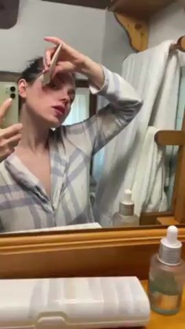A woman in a plaid pajama top stands in a bathroom, reflected in a mirror. She holds a cell phone in her left hand to record herself. With her right hand, she applies small drops of a white substance from a thin applicator onto her face. The bathroom includes a towel rack with a white towel, bottles on the counter, and a wooden shelf.
