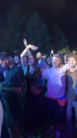 The video shows a crowd of people at an outdoor event at night. The foreground features several individuals dancing and interacting. A woman in a dark dress and green top has her arm raised, holding what appears to be a bill. A man in a white t-shirt and a cap stands next to her with his arms outstretched, and another man with curly hair is beside him, with his arm around the first man. The lighting is dim and colorful, with purple and blue hues dominating. In the background, more people are visible, along with trees silhouetted against the dark sky. The atmosphere appears festive and energetic, suggesting a concert or music festival.