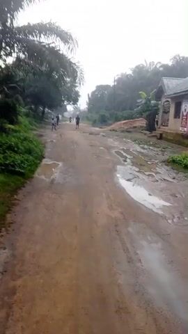 The video shows a muddy dirt road with puddles of water. There are buildings and trees on either side of the road. Several people and a motorcycle are seen moving along the road in the distance. The sky is overcast. The camera appears to be moving forward along the road.