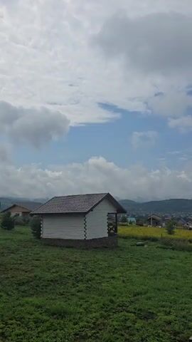 The video captures a pastoral landscape under a partly cloudy sky. In the foreground, a small, white wooden house with a dark-tiled roof sits on a lush green grassy hill. The house has a stone foundation and a covered porch area. Beyond the house, a village or town is visible, nestled among rolling hills that recede into the distance. The hills are covered in green foliage, and some areas appear to be cultivated with yellow flowers. The sky is a mix of blue sky and large, white, fluffy clouds, with some areas of light haze or mist around the distant hills. The overall scene suggests a rural or semi-rural setting, possibly in a mountainous region, on a day with dynamic weather conditions. There are no people or animals visible in the frame.