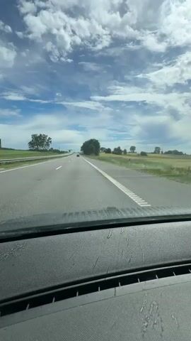 A first-person perspective video captures a view from inside a car driving on a highway on a sunny day with scattered clouds. The windshield is visible in the foreground, with the road and surrounding landscape stretching out ahead. As the video progresses, water appears on the windshield, and the windshield wipers are activated, smearing the water and obscuring the view of the road and landscape. The wipers move across the windshield multiple times, clearing some of the water but leaving streaks. The surrounding environment appears to be rural, with fields and trees lining the highway. There are a few other cars visible on the road in the distance. The sky is blue with white, puffy clouds. The video ends with the wipers still in motion, smearing water on the windshield.