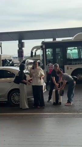 Four people are gathered around the open trunk of a silver sedan. A white shuttle bus is parked behind them. The person on the far left, wearing a black shirt and light-colored pants, stands facing the car, holding a black handbag over her left shoulder. To her right, a person in a white t-shirt and dark pants is looking at the car's trunk. A man in a dark blue t-shirt and dark pants stands to the right of the person in the white t-shirt, with his left hand raised. In front of the man, a person in a striped shirt and blue jeans is bending over, attending to a white suitcase with black trim. The ground appears to be wet, suggesting it might have rained recently. The sky is overcast.