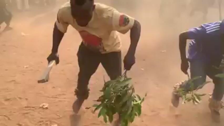 The video shows a group of people on a dusty, dirt ground. A man in the center wears a light-colored shirt, dark pants, and something wrapped around his ankles. He holds a flat object in his right hand and a leafy branch in his left. Two other men are visible near him, also holding leafy branches. A cloud of dust obscures the background, where a crowd of people appears to be watching.