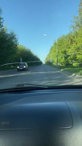 The video is filmed from inside a car, likely the driver's perspective, as it moves along a street lined with green trees on both sides. The road is paved and relatively clear of other traffic, with a single dark-colored car visible in the distance.  A white caution tape stretches across the road, obstructing the path. The sky above is clear and blue. Streetlights are visible along the road. The car's dashboard is visible in the foreground, showing the SRS airbag indicator.  The scene appears to be a residential or suburban area during daylight hours.