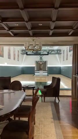 The video shows a basketball court with a glass wall in the foreground, separating it from a seating area. The basketball court has a wooden floor with green and white markings. There is a basketball hoop with a glass backboard and an orange rim. Behind the hoop, there are dark curtains and a statue of a person with a draped cloth. In the seating area, there is a table and two chairs, one of which is a brown armchair. The ceiling above the seating area has wooden beams and a chandelier. The lighting appears to be artificial.