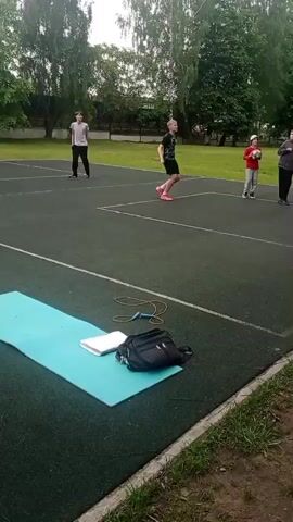 The video shows five children playing a game with a green basketball on a green outdoor basketball court. A woman is standing to the side, possibly supervising the children. A yoga mat, a jump rope, a notebook, and a small bag are laid out on the court, separate from where the children are playing. The court is surrounded by grass and several trees. The weather appears to be cloudy and mild.