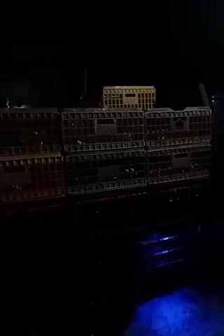 The video shows a stack of plastic crates in near darkness. The crates are stacked on what appears to be the bed of a truck or trailer, with blue light visible underneath. The crates vary in color, appearing to be dark gray, red, and yellow. The scene is mostly static, with only slight movement. The audio contains engine sounds.