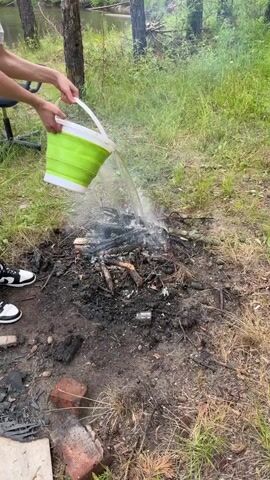 The video shows a close-up view of a campfire being extinguished with water. A person's hands are visible, holding a green collapsible bucket and pouring water onto the smoldering embers. Smoke billows from the fire as the water hits it. The campfire is situated on the ground in a natural setting with grass and trees in the background. There are also some discarded items, including a glass bottle and bricks, near the campfire.