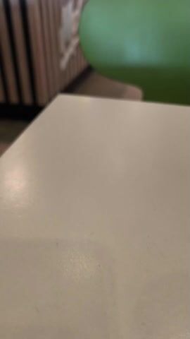 The camera slowly pans to the left, starting from a plain white table in the foreground. A bright green chair sits to the right of the table. The pan reveals a McCafe counter with a wooden slat design and a white McCafe logo. The background shows parts of the McDonald's restaurant, including the counter with display cases of food and a person walking by. The floor is tiled.