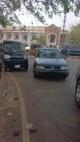 The video shows a street scene in what appears to be an urban area. Several cars are parked along the side of the road, including a dark blue Volkswagen Golf and a black van. A man in a checkered shirt and black pants walks along the sidewalk beside the parked cars. Other people are visible in the background, some riding on motorcycles. The road is paved but has debris scattered on it. In the background, there is a building with a light-colored exterior and several windows. Trees overhang the street, partially obscuring the sky.  A yellow three-wheeled vehicle is also seen in the background, along with other vehicles parked nearby. The overall atmosphere suggests a normal day in a populated area.