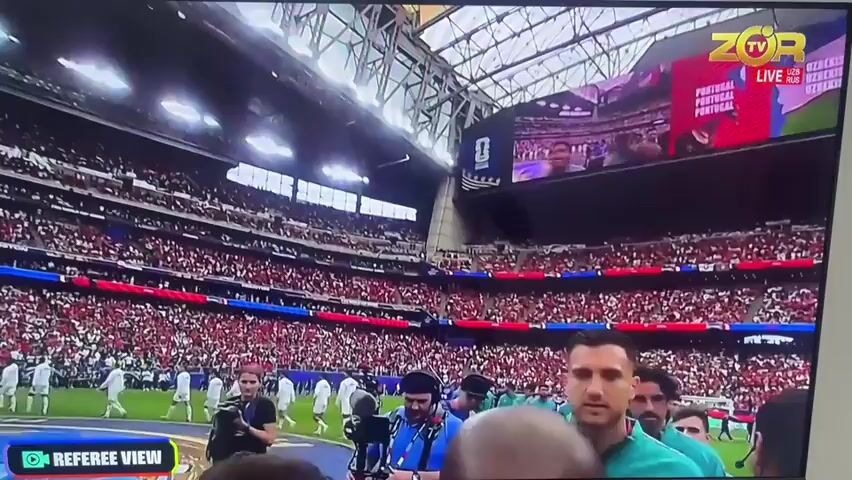 The media file shows a television screen displaying a sporting event. In the foreground, several people are visible, some wearing teal jerseys. Their expressions are varied, suggesting a range of emotions. The background depicts a crowded stadium with spectators in red and blue seating. Further back, there are individuals on a field, some holding cameras or equipment, implying a media presence.

The scene then shifts to a wider shot of the stadium's interior, focusing on the intricate metal structure of the roof. Hanging from the ceiling are American and Texan flags, along with several banners, one of which appears to be a logo with the text "ZOR TV LIVE UZB RUS". Another banner at the bottom of the screen reads "REFEREE VIEW".

The overall context suggests a live broadcast of a sporting event, possibly in the United States given the flags. The presence of "ZOR TV" indicates a broadcast from Uzbekistan. The event itself is not explicitly clear from the images, but the presence of athletes and a referee viewer designation hints at a competitive game. The atmosphere seems charged, with the crowds suggesting a significant event. There is no indication of the time of day or weather conditions.