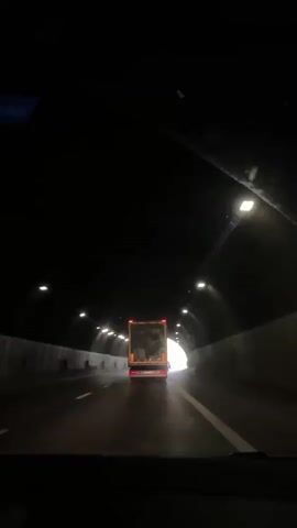 The video captures a view from inside a vehicle, driving through a tunnel. The primary object in focus is a truck ahead, its rear illuminated by the bright light emanating from the tunnel's exit. The tunnel itself is a concrete structure with a series of lights lining its ceiling, creating a dramatic contrast between the dark interior and the approaching daylight. The road surface appears wet, suggesting recent rain or damp conditions.

As the vehicle progresses, the tunnel opens up to reveal a scenic view. Lush green hillsides dotted with trees form the backdrop, bathed in the soft light of what appears to be daytime, likely late morning or early afternoon given the brightness. A clear blue sky with scattered clouds is visible above the hills. There are some road signs and utility poles visible on the right side of the road as the vehicle exits the tunnel.

The overall mood is one of transition and anticipation, moving from the enclosed darkness of the tunnel to the open, sunlit landscape. The video does not appear to contain any people within the frame besides the implied presence of the driver and any passengers within the vehicle from which the footage is recorded. The activity is simply travel through a tunnel and emerging into daylight. No text is visible in the video. The location context provided is Dolna Vasilitsa, Bulgaria.