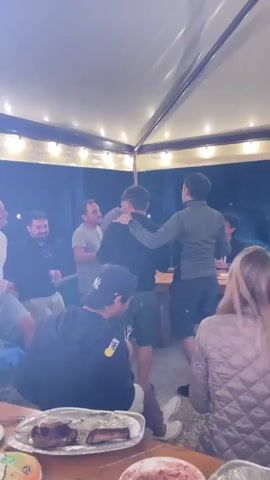 A group of people are gathered outdoors under a canopy with string lights. In the foreground, a person wearing a black hoodie and a New York Yankees cap is seated and holding a smartphone. Another person in a quilted jacket is seated to the right. In the middle ground, several men are standing. One man in a grey t-shirt with his arm in a cast is speaking animatedly, gesturing with his free hand. Two men in the center are interacting, with one man with dark hair and a beard appearing to embrace the other man with dark hair and no beard, who is wearing a black jacket. To the right, a man in a green jacket is standing with his back to the camera. A table with food, including what appears to be ribs or steaks on a platter, is in the foreground. The overall impression is a casual social gathering.