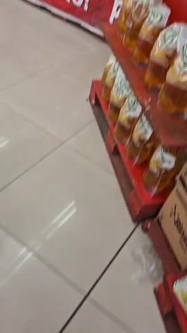 The video begins with a close-up view of shelves stocked with bottles of cooking oil in clear plastic packaging, each with a green and white label. The bottles are arranged in rows on a red wooden display. The camera then pans to the right, revealing a large red banner with white text that reads "SUPER PROMO!". Behind the banner, more shelves are visible, stocked with various household products, including detergent and fabric softener, in colorful packaging. The store environment appears to be a supermarket or grocery store, with tiled flooring. The camera continues to pan, showing aisles of shelves filled with packaged food items like snacks and biscuits. Further down the aisle, a person wearing a black jacket and white pants is seen browsing the shelves, with a red shopping basket near them. The video ends by showing a shopping cart in the foreground.