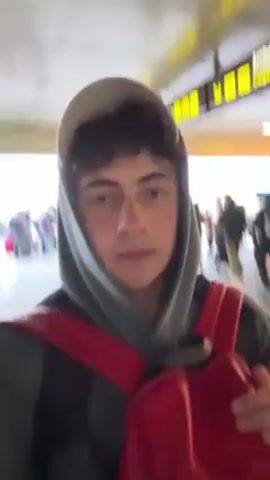 Here's a description of the video:

The video shows a young man wearing a grey hoodie, a backwards white baseball cap, and a red backpack, likely at an airport. He appears to be filming himself as he walks through a busy area. Other people with luggage are visible in the background. He looks directly at the camera and doesn't speak. He walks into a building or hallway with glass walls. The light changes as he moves inside.
