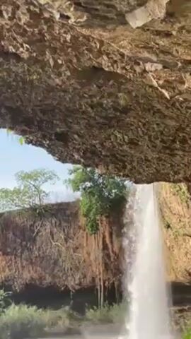 Viewed from under a rocky overhang, a tall, narrow waterfall cascades into a shallow pool, creating a misty spray. The overhang, composed of textured, light-brown rock, dominates the foreground. Beyond the waterfall, a brown, earth cliff is topped with green trees and trailing vines. A clear sky is visible above the trees, contrasting with the textured rock and flowing water.