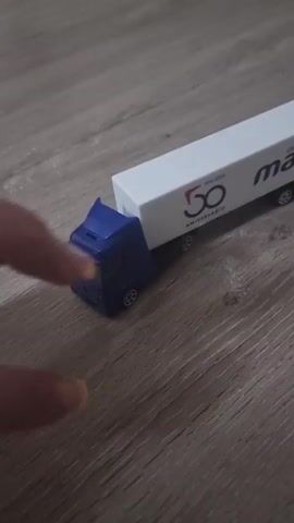 A close-up video shows a person's hand interacting with miniature toy vehicles on a light-colored wooden floor. The hand, which has a tattoo on one finger and yellow nail polish on another, is first seen detaching a small blue toy truck cab from a white toy trailer. The trailer features the text "GRUPO mazo" with a red arrow logo, and "50 ANIVERSARIO" below a large "50" emblem. After a brief separation, the hand re-attaches the blue truck cab to the white trailer. The hand then pushes the assembled toy truck and trailer forward across the wooden surface, moving from left to right across the frame. There are no visual cues within the video to specifically indicate the provided location context of Xàtiva, Spain.