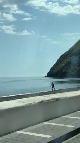 A wide shot captures a serene coastal scene in Lipari, Italy, on a bright, partly cloudy day. In the foreground, a white concrete wall runs horizontally, separating the viewer's perspective from a pebble beach and the calm, blue sea. A lone individual, silhouetted against the bright sky and water, stands on the edge of the beach, actively throwing an object into the sea. Their body is in motion, with one arm extended forward from a bent stance, indicating the throwing action.

The sea stretches out calmly to the horizon, reflecting the soft blue of the sky and scattered white clouds. On the right, a lush green hill or mountain rises from the coastline. In the far distance, across the expanse of water, two distinct mountain peaks are visible, suggesting other islands or distant landmasses. The overall atmosphere is peaceful and clear. No text is visible in the frame.