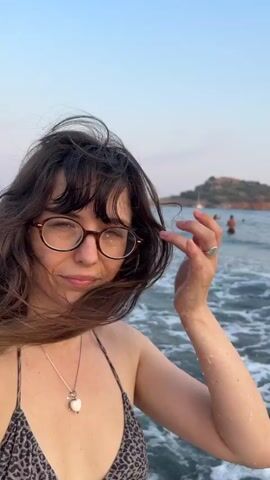 A woman with long, dark hair and glasses is standing in the ocean. She is wearing a leopard-print bikini and a necklace with a white pendant. The water is choppy and there are waves. In the background, there is a rocky island and a clear sky. The woman is looking at the camera and smiling slightly. She raises her hand to her face and touches her hair. The video is short and appears to be a selfie taken by the woman.