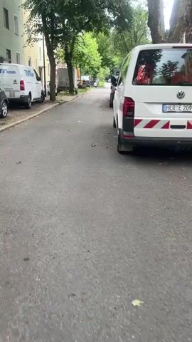 A low-angle shot on a paved street shows a white van with "EKW" on its side parked on the right, followed by a black sedan and a small pickup truck further down the street. Another white van is parked on the left. The street is lined with trees and residential buildings, and there are scattered leaves on the asphalt. The video pans slightly to the right, maintaining a clear view of the parked vehicles and the street ahead.