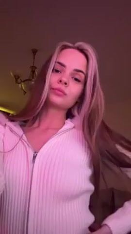 A young woman stands indoors, filmed from a low angle. She is fair-skinned with long brown hair, highlighted with blonde streaks near her face. She wears a white ribbed zipper hoodie. The lighting in the room is pink. In the background, a chandelier and a piece of furniture are visible. The woman looks into the camera and then glances to the side, holding a pose. Music is playing in the background.