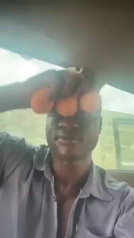 A person is shown in a car, holding three brown eggs. He puts one of the eggs in his mouth. He then places the other two eggs on his forehead, balancing them above his eyes. The video is shot from the perspective of the person in the car, looking directly at their face.  The person is wearing a light blue shirt and a cap. The background shows the interior of the car and glimpses of the outside through the windows.