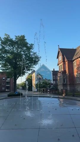 A short video clip showcases a public square with a fountain in the foreground. The fountain features multiple jets of water arcing upwards, creating a dynamic visual effect. The square is paved with large, light-colored tiles, some of which are wet and reflective from the fountain's spray.

In the background, a vibrant mural adorns the side of a building. The mural depicts a scenic landscape with a cloudy blue sky, rolling hills, and possibly a structure or figures within the scene, though the details are somewhat abstract. To the left of the mural, a large, leafy green tree stands prominently, its branches reaching towards the sky.

Surrounding the square are buildings with traditional European architecture, characterized by red brickwork and pitched roofs. The buildings appear to be residential or commercial properties. A street is visible behind the fountain, with a single blue traffic sign visible on the left.

The time of day appears to be early morning or late afternoon, suggested by the clear, bright blue sky and the gentle lighting. The weather seems to be fair and pleasant. The overall atmosphere is serene and calm, with the gentle sound of the fountain likely contributing to the peaceful ambiance. There are no people visible in the clip.

The location is identified as Vejle, Denmark. No discernible text is visible in the video clip.
