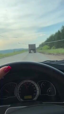 The video begins with a point-of-view shot from inside a car, looking out the windshield. The driver's hands, with red painted nails, are visible on the steering wheel. The car's dashboard displays a speedometer reading of approximately 110 km/h and an odometer reading of 390336. The road ahead is asphalt with a yellow line marking the lane. A large truck is visible in the distance, also on the road. The sky is cloudy with some patches of blue. The landscape on either side of the road consists of green fields and trees. The camera then pans to the right, showing the side mirror reflecting a car following behind. The sun is visible in the sky, partially obscured by clouds. The scenery continues to be rural, with fields and distant hills. The video ends with the camera focused on the side mirror, showing the car behind them.