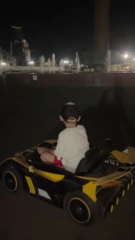 A young child, wearing a white t-shirt, red shorts, and a baseball cap, sits in the driver's seat of a black and yellow toy car at night. The child's back is to the camera as they hold the steering wheel. The toy car is equipped with a spoiler on the rear and has the markings "M76.4" on the back. The child is holding onto a black backpack that is placed behind them in the car. The background is dark, suggesting it is nighttime. In the distance, there are illuminated fountains and structures, possibly part of a park or city square. The ground appears to be paved. The lighting suggests artificial illumination from the city or the fountains.