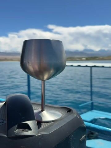 Here's a description of the video:

The video shows a metal wine glass sitting on the edge of a boat. The boat is moving on a body of water, with mountains and a cloudy sky visible in the background. The boat has blue railings and a small ladder. The water is a clear blue color. The sun shines brightly.
