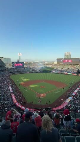 This video begins with a wide shot of a baseball stadium filled with spectators. The camera then zooms in on the baseball field, focusing on players in uniform. The scene is bathed in the warm light of what appears to be late afternoon or early evening. Smoke or fog drifts across the field, obscuring some of the outfield. The stadium features large screens displaying the "Angels" logo. A few fireworks are visible in the upper left corner of the frame. The crowd is visible in the stands, some wearing red. The video is shot from a high vantage point, looking down onto the field.