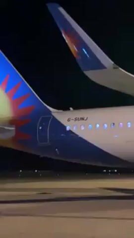 The video shows the side of a parked Jet2holidays airplane at night. The plane is white with a blue stripe along the fuselage.  "Holidays you can trust" is written in red above the windows.  The engine has "Jet2holidays" printed on it in orange letters. A small vehicle and personnel can be seen under the wing.