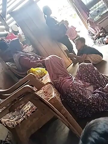 The video shows an interior scene with several people. In the foreground, two individuals are seated in chairs facing away from the camera. One person is wearing a patterned headscarf and a long-sleeved patterned dress. The other person is wearing a maroon headscarf and a maroon top. A yellow bag is placed on the lap of the person in the maroon headscarf. In the mid-ground, a young boy is crouched down, holding a mobile phone and interacting with an adult who is partially visible. Behind them, another person, possibly a child, stands with their back to the camera. Further in the background, another individual wearing a pink headscarf is seated. The environment appears to be a room with wooden furniture, including chairs and what looks like a table or counter. Sunlight is streaming in from an opening, casting light and shadows across the scene. The overall impression is of a casual, domestic gathering.