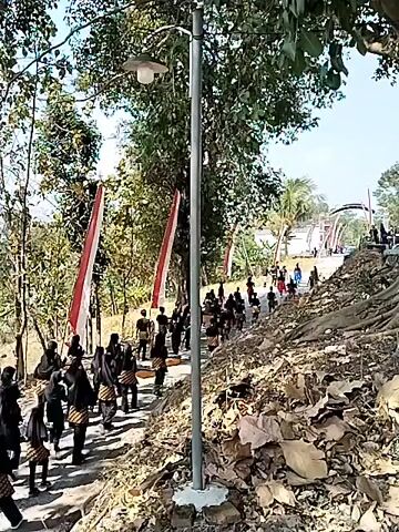 A group of people in traditional clothing, some carrying baskets, walk along a dirt path lined with trees and foliage. Red and white flags are visible, along with a banner that reads "KARANG TARUNA". A lamppost stands near the path. The ground is uneven and covered with dry leaves and exposed roots. The sun is shining, casting shadows.