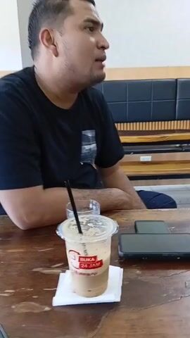The video shows a man sitting at a wooden table with a disposable plastic cup of iced coffee, a glass of water, and two smartphones. The cup has a logo that reads "BUKA 24 JAM" above "jalahkuhipi". He is wearing a black t-shirt. The man is speaking. Behind him is a black leather bench with a wooden back and base. The camera is still and close-up, focusing on the man and the items on the table.