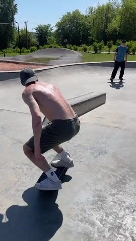 The video shows two young men at a skate park on a sunny day. In the foreground, a shirtless young man with a baseball cap and shorts is skateboarding. He is wearing white sneakers and is in the process of performing a trick on a slanted metal ramp. His shadow is cast on the concrete ground. In the background, another young man wearing a blue shirt and dark pants is standing on his skateboard. The skate park features concrete ramps and curves, surrounded by green grass and trees. The sky is clear and blue.