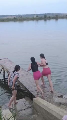 Three women stand on a wooden pier extending over a body of water. The woman on the far left wears a floral print shirt and denim shorts. The two women on the right wear swimsuits. The woman in the middle wears a black top and pink shorts, and the woman on the right wears a black bikini top and pink shorts. The two women on the right jump into the water. The woman in the middle jumps first, followed closely by the woman on the right. The woman in the floral print shirt walks further onto the pier. The water is calm and the sky is overcast. The pier is old and weathered. The surrounding landscape is green and hilly.